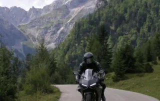 Motorradfahrer auf einer kurvenreichen Straße in den Alpen, umgeben von beeindruckenden Bergen und üppiger Natur.