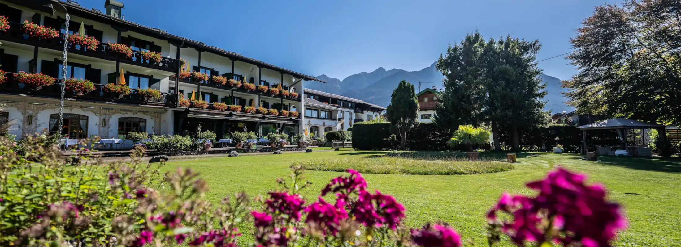 Der Garten des familienfreundlichen Hotels mit bunten Blumen, Sonnenschirmen und Blick auf die umliegenden Berge in Bayern.
