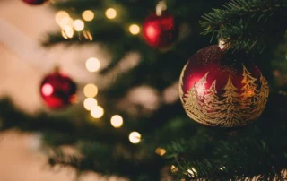 Rote Weihnachtskugel mit goldener Baumgestaltung hängt am Weihnachtsbaum, umgeben von weihnachtlicher Beleuchtung und Tannenzweigen.