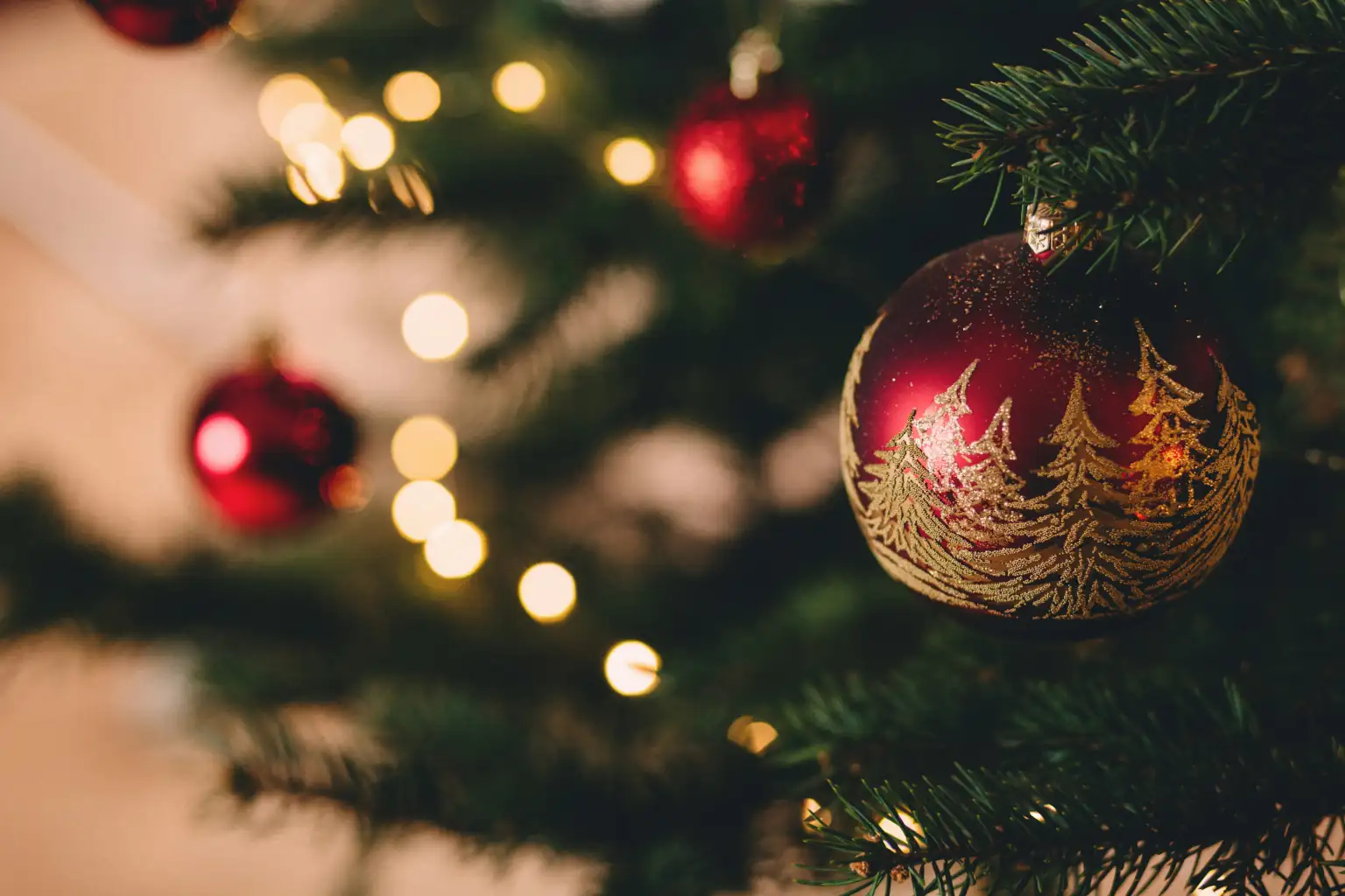 Rote Weihnachtskugel mit goldener Baumgestaltung hängt am Weihnachtsbaum, umgeben von weihnachtlicher Beleuchtung und Tannenzweigen.